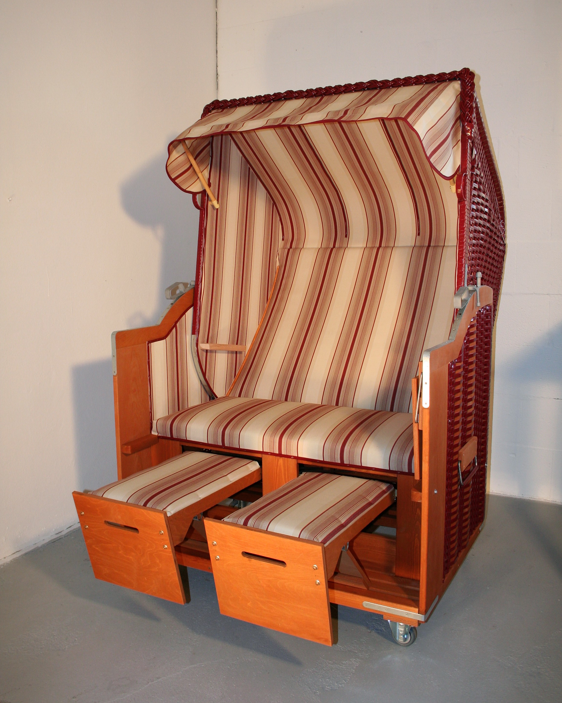 Der 2-Sitzer Strandkorb hat ein Geflecht aus PVC in der Farbe bordeaux mit einem gestreiften Stoffdesign in beige-rot.